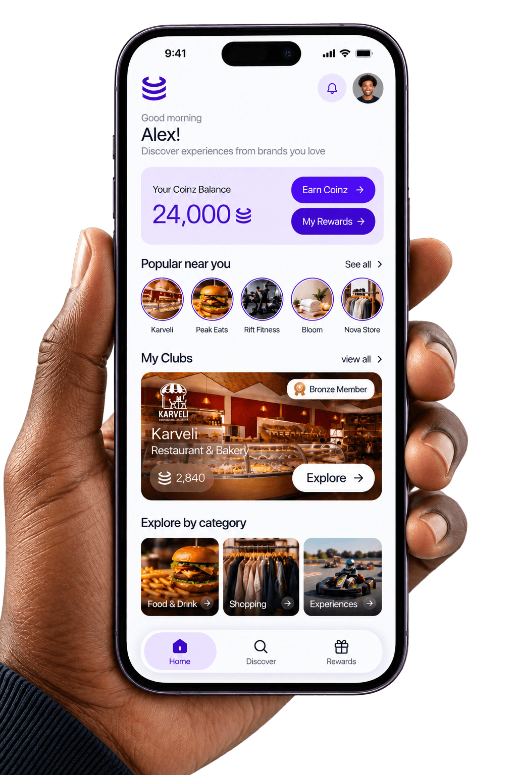 A hand holding a phone showing the Coinz app home screen, with a balance of 24,000 Coinz and places nearby